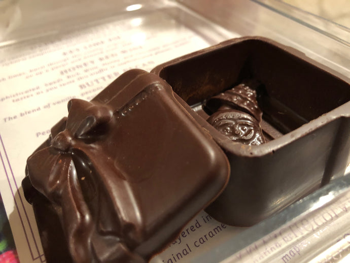 A dark chocolate gift box with a clear window, showing the interior with a few chocolate pieces. The exterior has a decorative bow and text indicating it is a 2 1/2 inch by 2 1/2 inch size.